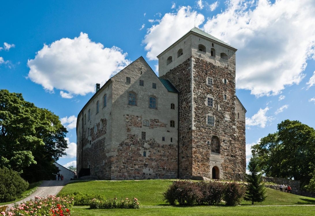 En velbevart middelalderfestning eller borg i stein med et fremtredende firkantet tårn, bygget med en kombinasjon av stein og hvitkalkede vegger. Strukturen ligger på en liten høyde omgitt av anlagte områder med trær og blomsterbed. Bildet er tatt på en lys dag med blå himmel og hvite skyer.