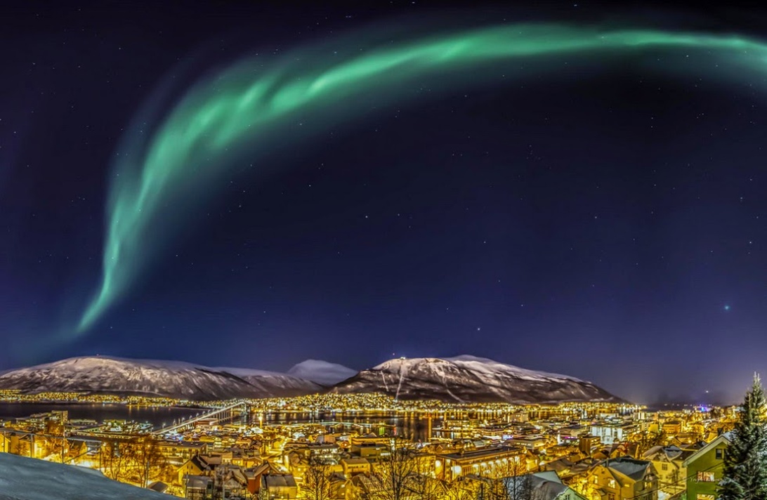 En fantastisk nattlig byscene, sannsynligvis Tromsø, Norge, med spektakulært grønt nordlys som danser over den mørke himmelen. Snødekte fjell rammer inn den sterkt opplyste byen nedenfor, og skaper en klassisk norsk vinterscene. Dette illustrerer en populær vinterattraksjon for turister i Norge.